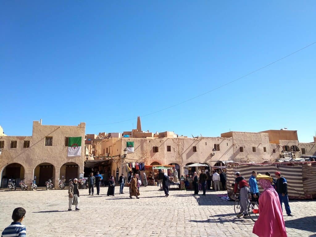 The Labyrinths of the M'zab Valley: In Search of the Cyclopes of Ghardaïa 2 Ghardaïa central market square in Algeria's M'zab Valley, with arcaded ochre ksar buildings and the pyramidal Ibadi minaret of the Mozabite Berber citadel rising on the hilltop — a UNESCO World Heritage Saharan pentapolis town.