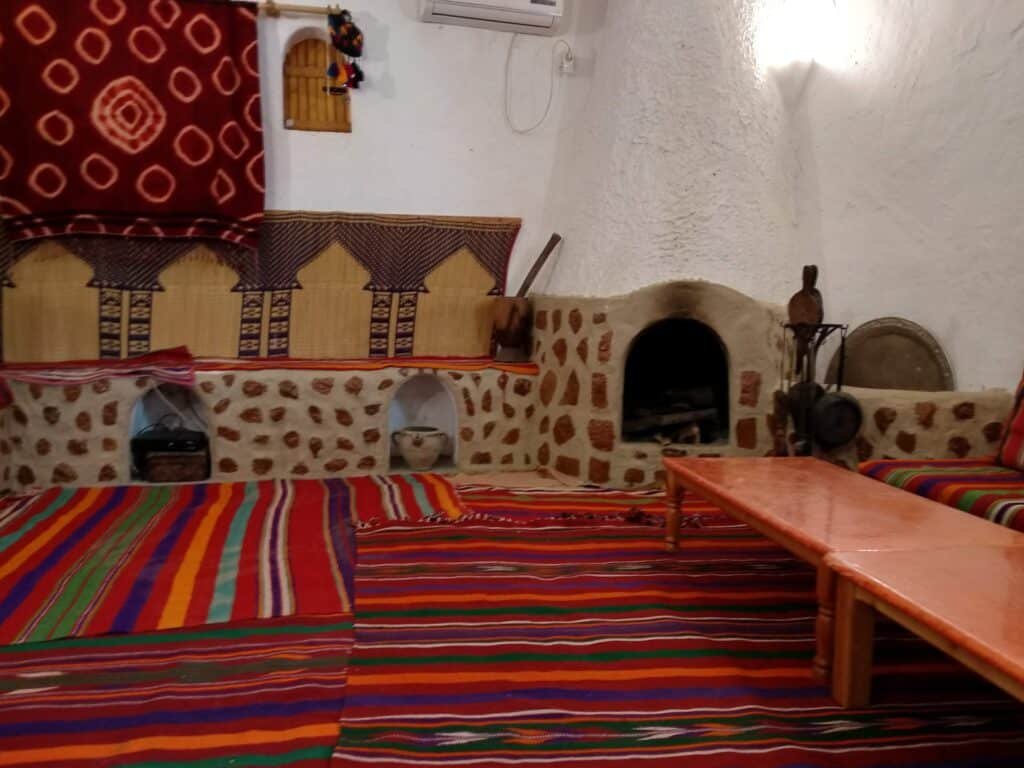 The Labyrinths of the M'zab Valley: In Search of the Cyclopes of Ghardaïa 4 Traditional Mozabite Berber interior in Ghardaïa, M'zab Valley — a whitewashed living room with a stone hearth, wall niches, woven palm-fibre mats and striped rugs on the floor, showing the vernacular M'zab architecture and Ibadi domestic life of Algeria's Sahara.